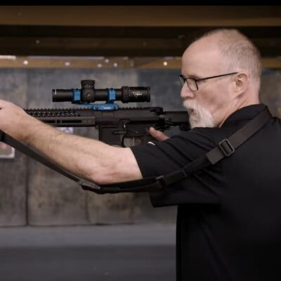 Patrick Kelley looking down the scope of a rifle with the sling over his shoulder