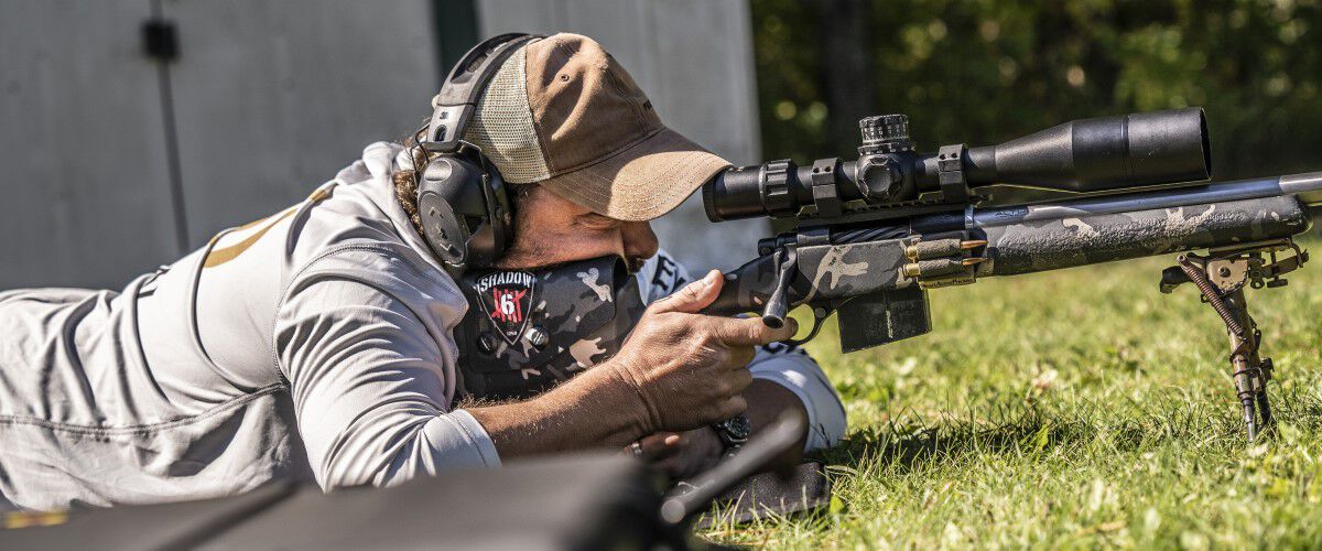 Jim Gilliland laying on the grass looking down the scope of a rifle on a bipod