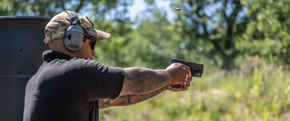 guy standing next to a barrel and pointing a pistol